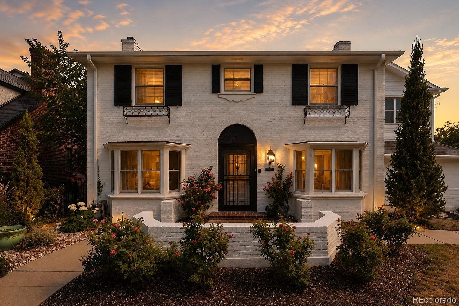 Twilight view of white-brick Colonial framed by rose gardens